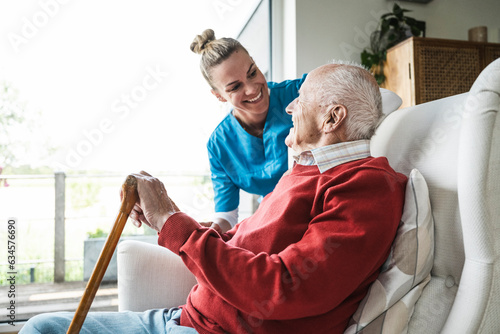 Happy nurse taking care of senior man sitting in armchair at home photo