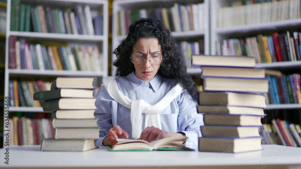 Tired confused female student studying by reading books in campus library. Exhausted girl learning on examination period at the university. Overworked applicant spreads her hands and looks at camera Tired confused female student studying by reading books in campus library. Exhausted girl learning on examination period at the university. Overworked applicant spreads her hands and looks at camera