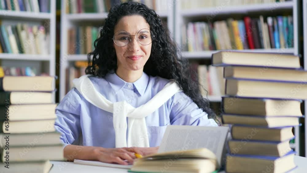 Portrait of smiling female student sitting at desk for learning in campus library space. Stylish brunette girl in glasses looks at the camera. Applicant prepares for university exams in the classroom Portrait of smiling female student sitting at desk for learning in campus library space. Stylish brunette girl in glasses looks at the camera. Applicant prepares for university exams in the classroom