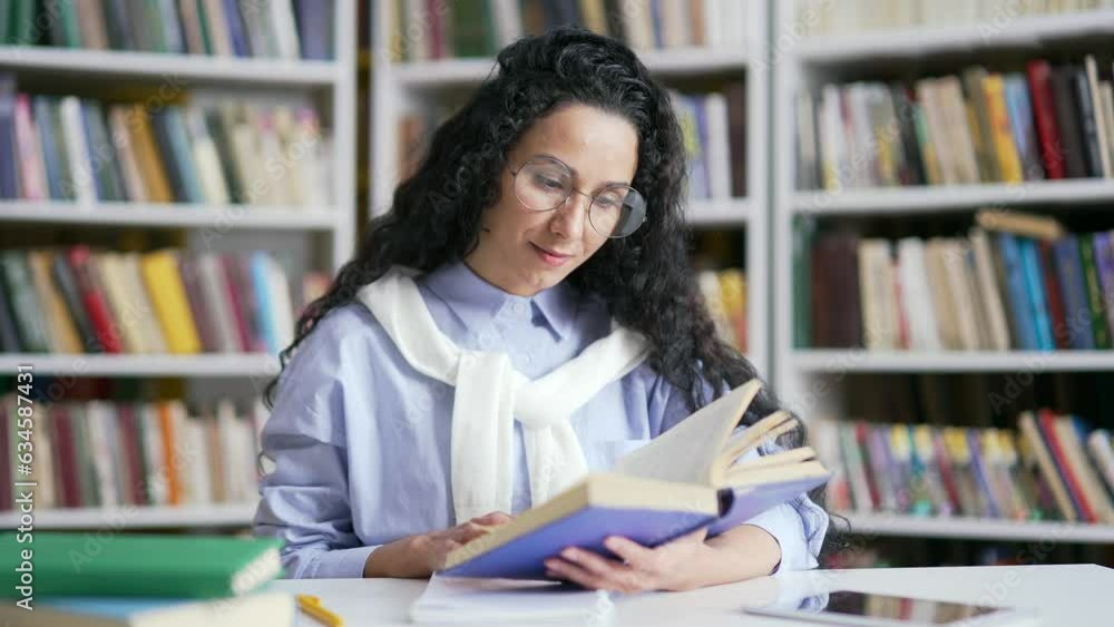 Smiling female student learning reading a book in campus library space. The girl is studying for the examination period at the university in the classroom. Applicant prepares for the entrance exams Smiling female student learning reading a book in campus library space. The girl is studying for the examination period at the university in the classroom. Applicant prepares for the entrance exams
