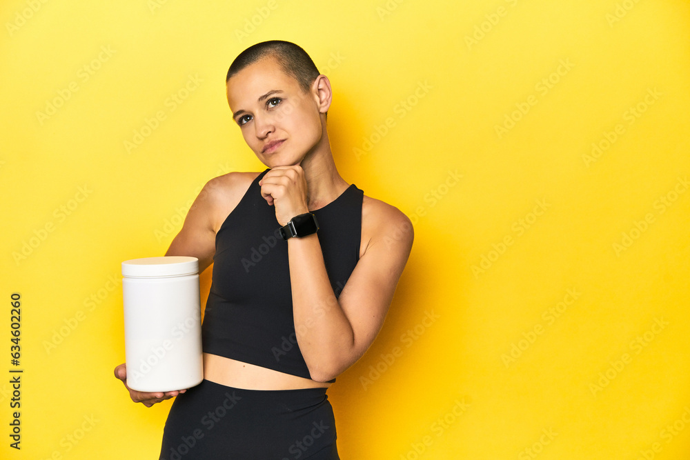 Sportswoman with protein shake, yellow studio background looking sideways with doubtful and skeptical expression. Sportswoman with protein shake, yellow studio background looking sideways with doubtful and skeptical expression.