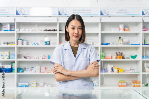 Happy handsome asian female pharmacist wearing lab coat standing with arms crossed and looking at camera, She feels good, trustworthy and proud of his work in the pharmacy drugstore.