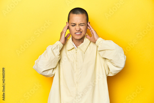 Young woman in jacket  yellow studio backdrop covering ears with fingers  stressed and desperate by a loudly ambient.