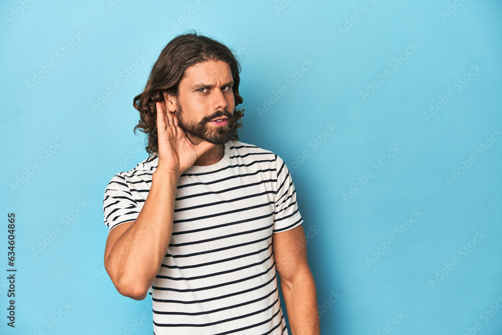 Bearded man in a striped shirt, blue backdrop trying to listening a gossip. Bearded man in a striped shirt, blue backdrop trying to listening a gossip.