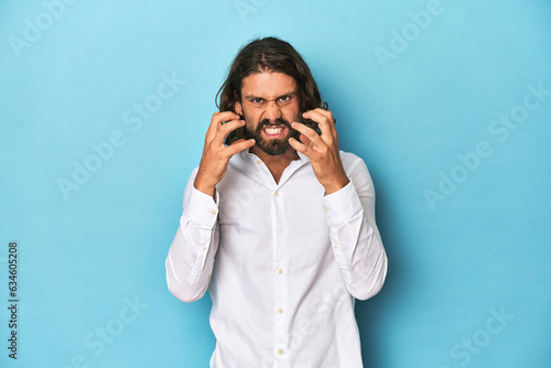 Bearded man in a white shirt, blue backdrop upset screaming with tense hands.
