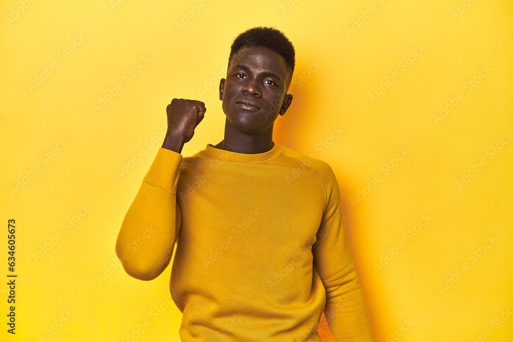 Stylish young African man on vibrant yellow studio background, showing fist to camera, aggressive facial expression. Stylish young African man on vibrant yellow studio background, showing fist to camera, aggressive facial expression.