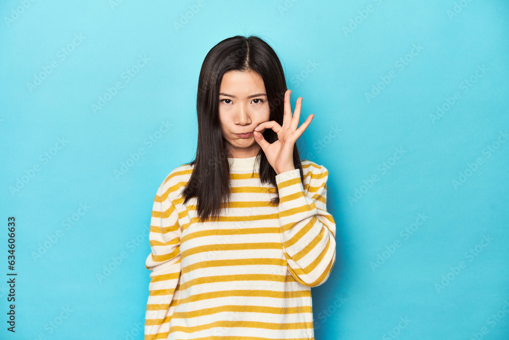 Asian woman in striped yellow sweater, with fingers on lips keeping a secret. Asian woman in striped yellow sweater, with fingers on lips keeping a secret.