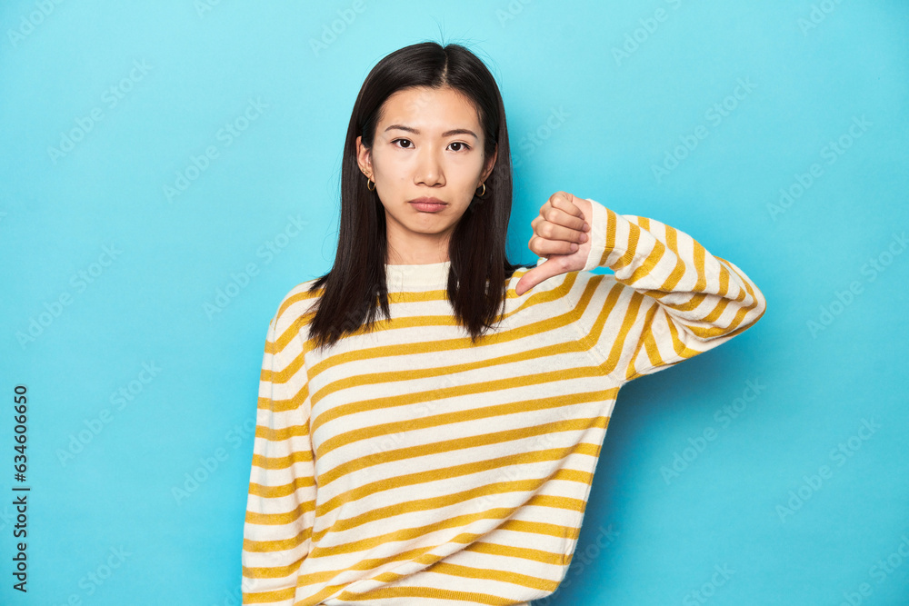 Fototapeta premium Asian woman in striped yellow sweater, showing a dislike gesture, thumbs down. Disagreement concept.