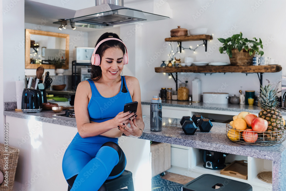 young latin woman resting after workout at home using mobile phone and headphones at kitchen in Home in Mexico Latin America, hispanic female in wellness concept