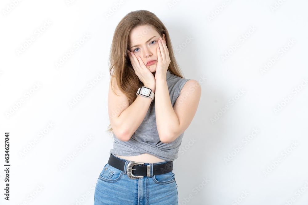 Young beautiful girl wearing grey T-shirt keeps hands on cheeks has bored displeased expression. Stressed hopeless model Young beautiful girl wearing grey T-shirt keeps hands on cheeks has bored displeased expression. Stressed hopeless model
