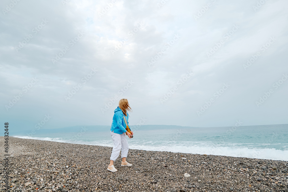 Attractive blond woman in light blue hoodie walking on winter beach. Traveling. Wellbeing and harmony concept. Unity with nature. Attractive blond woman in light blue hoodie walking on winter beach. Traveling. Wellbeing and harmony concept. Unity with nature.
