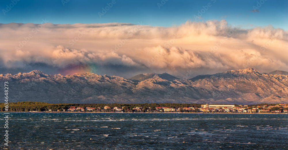 Vrsi, Croatia - Panoramic view of Vrsi municipality in Zadar county with Velebit mountains and Paklenica national park covered with warm golden colored clouds and rainbow in background at sunset