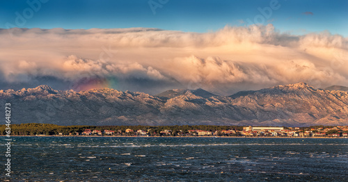 Obraz na płótnie Vrsi, Croatia - Panoramic view of Vrsi municipality in Zadar county with Velebit mountains and Paklenica national park covered with warm golden colored clouds and rainbow in background at sunset w salonie