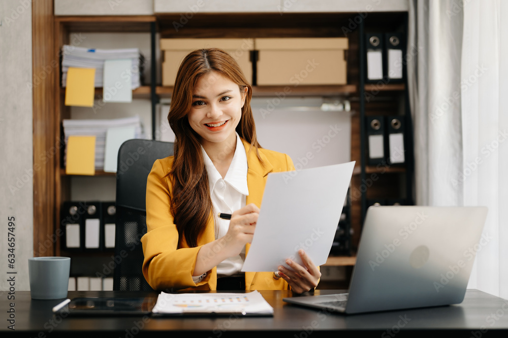 Smiling Asian woman talking on the phone with a customer Young positive female accountant using smartphone talking to team at her desk relaxing in office Smiling Asian woman talking on the phone with a customer Young positive female accountant using smartphone talking to team at her desk relaxing in office