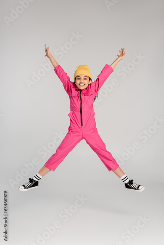 excited girl in trendy beanie hat and pink outfit jumping with outstretched hands on grey, have fun