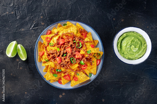 Nachos, Mexican food, tortilla chips with beef and fresh vegetables, top shot on a black slate background with limes and guacamole dip