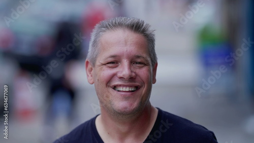 Joyful man smiling and laughing while standing in city street looking at camera. Portrait face close-up of a happy 40s person. Real life authentic people