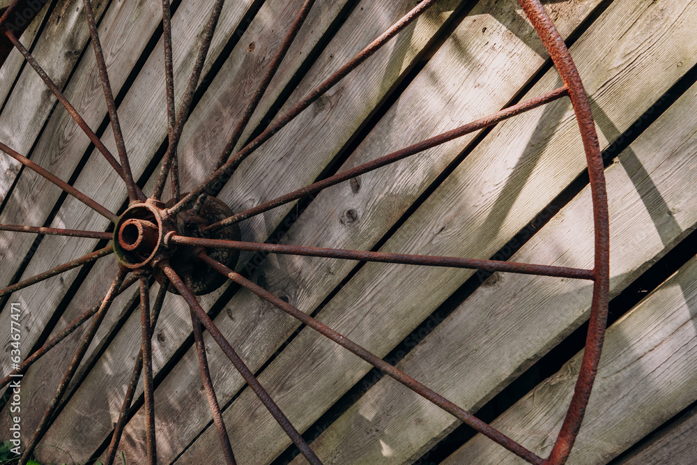An old rusty wheel from a large cart against the background of a dilapidated unpainted fence. Abandoned place of rural type with a rusty wagon wheel close-up An old rusty wheel from a large cart against the background of a dilapidated unpainted fence. Abandoned place of rural type with a rusty wagon wheel close-up
