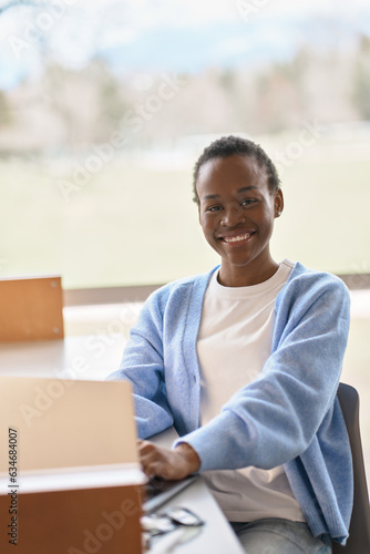 Smiling short-haired African Black girl student using laptop computer technology sitting at desk. Happy pretty young woman elearning looking at camera advertising hybrid work. Vertical portrait.
