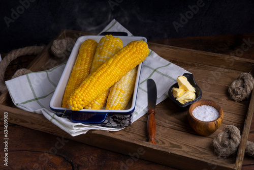 Boiled yellow corn in a ceramic mold, white kitchen towel, butter, vintage knife, salt in a salt shaker on an antique wooden tray.