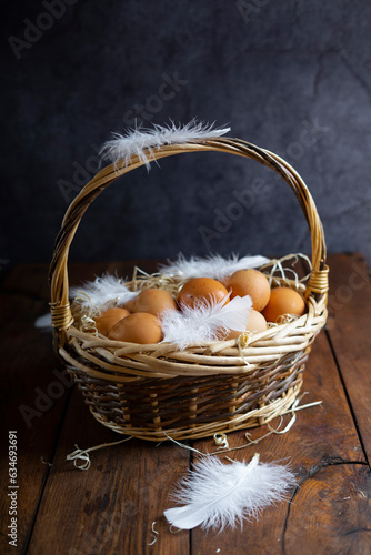 Vine basket filled with fresh chicken brown eggs, white feathers on a dark wooden background.