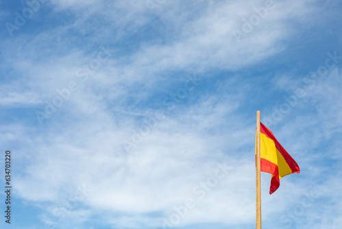 spanish flag on blue sky with white clouds and wooden mast.
