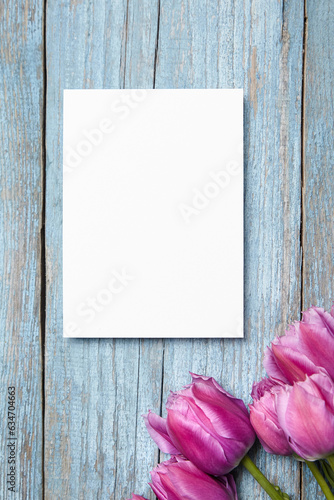 Blank invitation card mockup, white greeting card with purple tulip flower on a blue wooden background, flat lay. Top view of blank postcard with floral accents