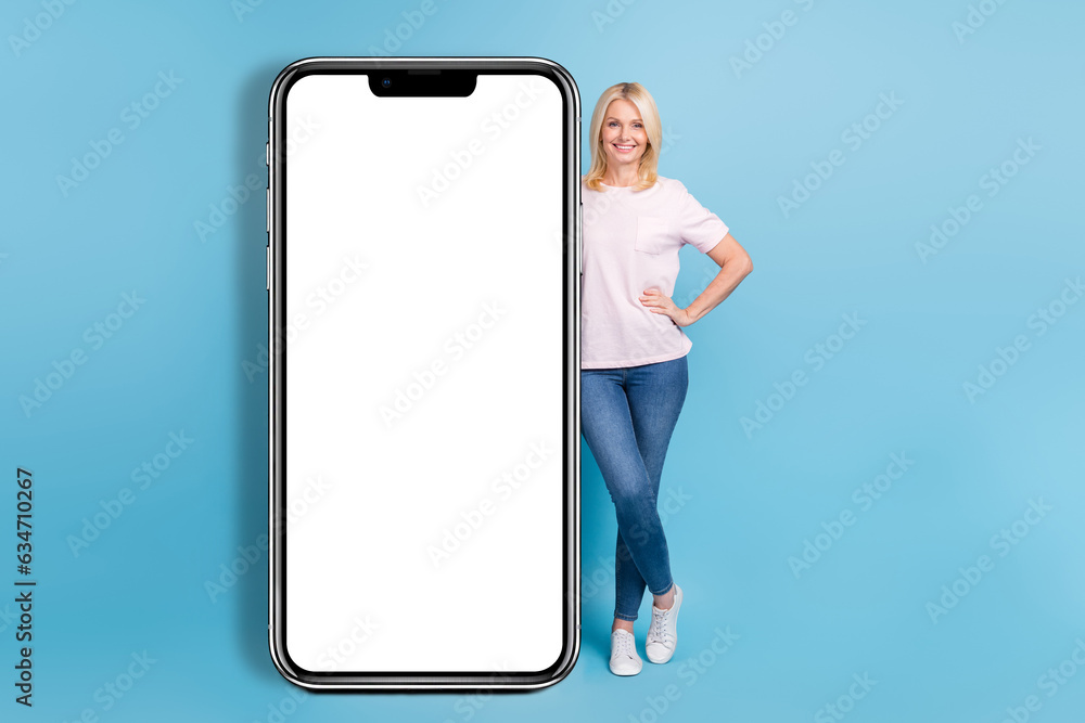 Full size photo of positive pensioner woman white t-shirt jeans sneakers stand near panel empty space isolated on blue color background Full size photo of positive pensioner woman white t-shirt jeans sneakers stand near panel empty space isolated on blue color background