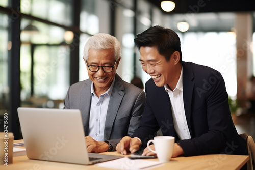 Mature boss manager mentor helping Asian employee working on laptop in office. Two happy professional coworkers discussing online plan at work. Diverse business people using computer and talking