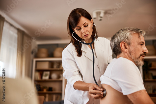 Female doctor holding stethoscope examining senior male patient sitting at home.