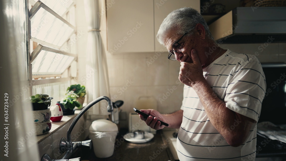 Pensive senior man looking at phone screen pondering dilemma and decision-making with a smartphone device while standing in home kitchen by window Pensive senior man looking at phone screen pondering dilemma and decision-making with a smartphone device while standing in home kitchen by window