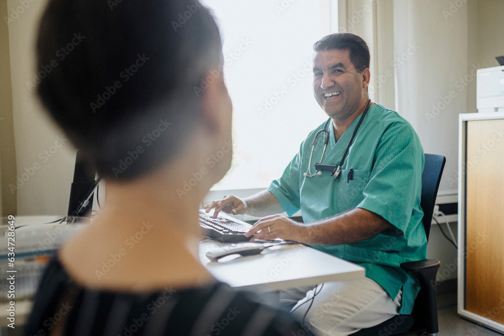 Happy male doctor looking at patient while using computer in clinic Happy male doctor looking at patient while using computer in clinic