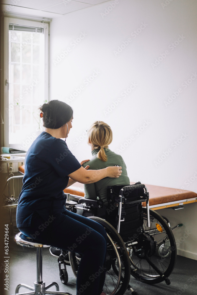 Side view of female nurse examining woman sitting on wheelchair in clinic Side view of female nurse examining woman sitting on wheelchair in clinic