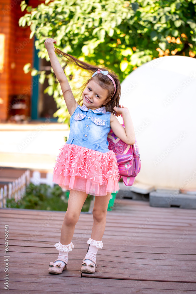 Cute little girl holding a pink backpack, playing in the yard. Beautiful little girl smiles in a smart dress. Preschool education, preparation for school. Cute little girl holding a pink backpack, playing in the yard. Beautiful little girl smiles in a smart dress. Preschool education, preparation for school.