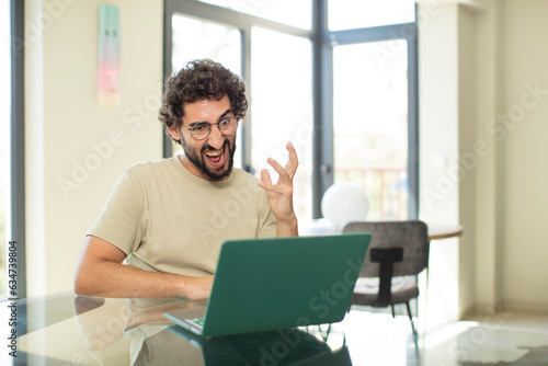 young adult bearded man with a laptop looking angry, annoyed and frustrated screaming wtf or what’s wrong with you