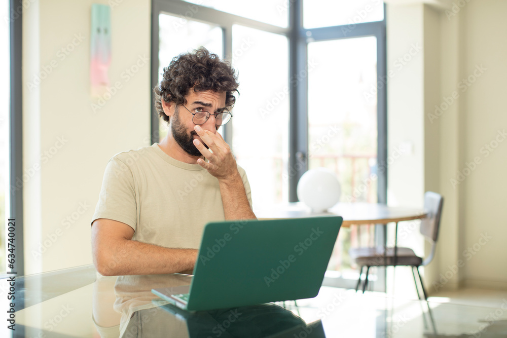 young adult bearded man with a laptop feeling disgusted, holding nose to avoid smelling a foul and unpleasant stench young adult bearded man with a laptop feeling disgusted, holding nose to avoid smelling a foul and unpleasant stench