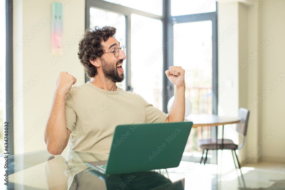 young adult bearded man with a laptop feeling happy, positive and successful, celebrating victory, achievements or good luck young adult bearded man with a laptop feeling happy, positive and successful, celebrating victory, achievements or good luck