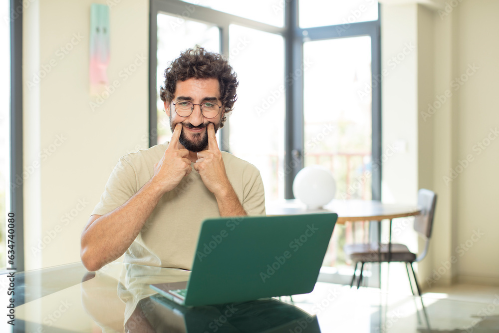 young adult bearded man with a laptop smiling confidently pointing to own fake smile young adult bearded man with a laptop smiling confidently pointing to own fake smile