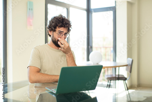 young adult bearded man with a laptop feeling disgusted, holding nose to avoid smelling a foul and unpleasant stench