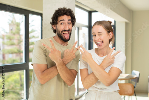 young adult couple smiling and looking happy, friendly and satisfied, gesturing victory or peace with both hands