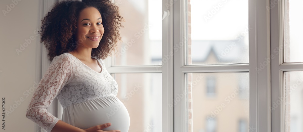 Young pregnant black woman smiling and looking at empty space enjoying her pregnancy near a window at home - obrazy, fototapety, plakaty 
