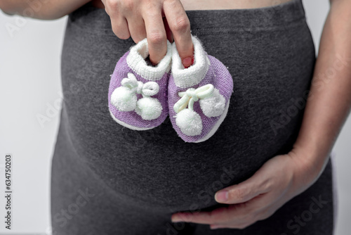 Close-up of pregnant woman touching her belly and holding small baby shoes on pregnant belly in the home for baby girl on the future concept.