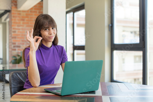 young pretty woman feeling happy, showing approval with okay gesture. desk laptop concept
