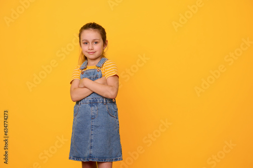 Beautiful little girl in blue denim overalls, smiles looking at camera, posing with arms folded over orange background