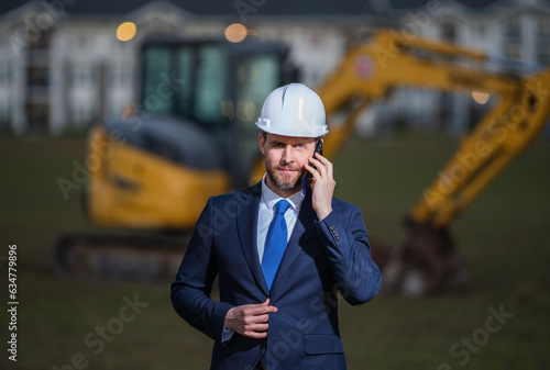 Construction owner near excavator. Confident construction owner in front of house. Architect, civil engineer. Man construction owner with a safety vest and hardhat at construction site.