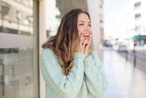 pretty hispanic woman feeling happy, excited and surprised, looking to the side with both hands on face