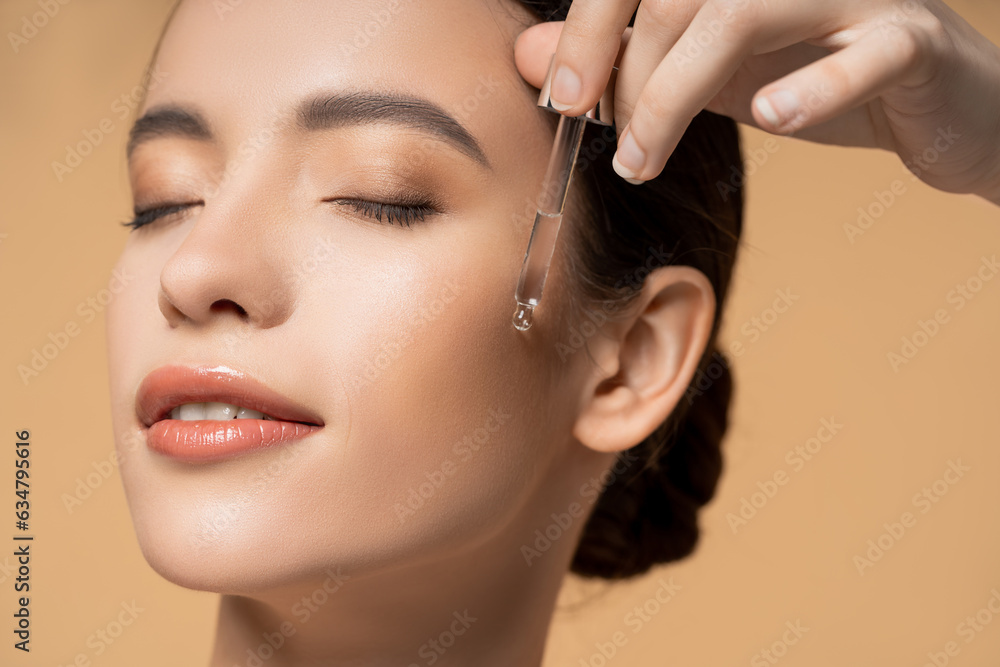 Close up view of pleased asian woman applying cosmetic serum  while standing isolated on beige