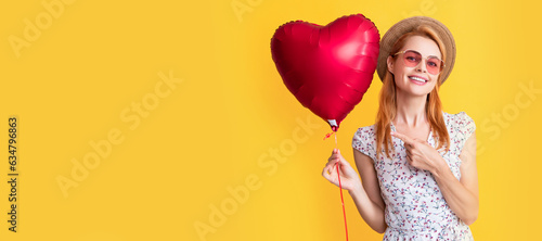 happy young woman in straw hat and sunglasses point finger love heart balloon on yellow. Woman isolated face portrait, banner with copy space background. © Olena