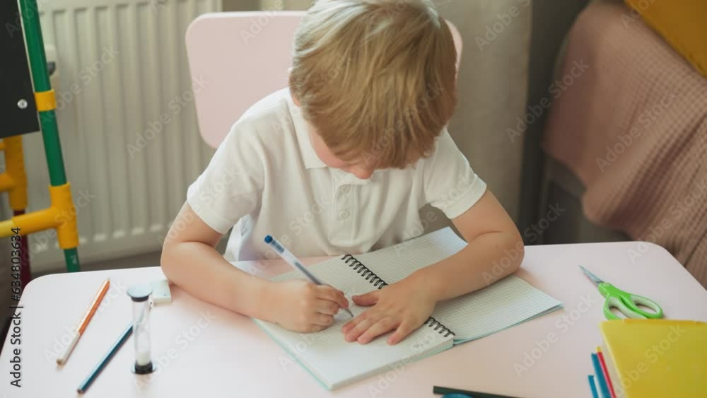 Little blond pupil draws spot with pen and wipes by finger in notebook in home classroom. Toddler boy does creative work in copybook in kindergarten