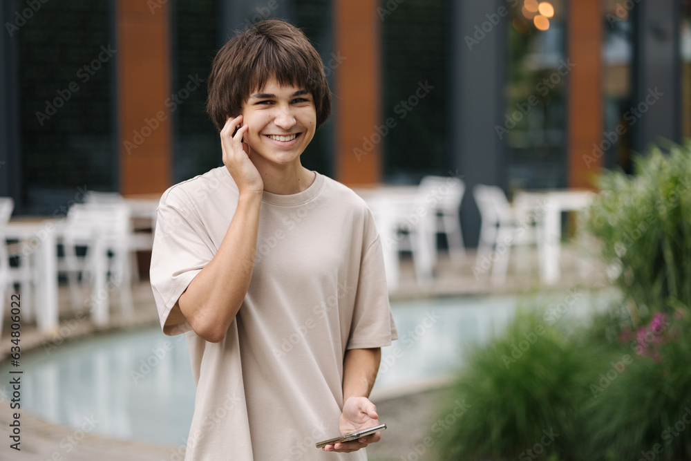 Happy smiled boy speak by wireless headphones and hold his smartphone in hands. Background of terrace in cafe